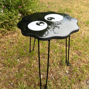 May include: Black side table with a black and white cartoon eye design. The table has a unique, irregular shape and is supported by four thin black metal legs.