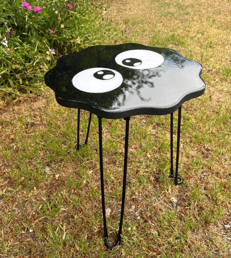 May include: Black side table with a black and white cartoon eye design. The table has a unique, irregular shape and is supported by four thin black metal legs.