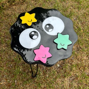 May include: A black resin table with a wavy edge, decorated with four colourful stars and two white eyes. The table has a black metal leg.
