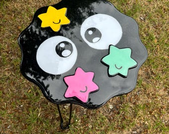 May include: A black resin table with a wavy edge, decorated with four colorful stars and two white eyes. The table has a black metal leg.