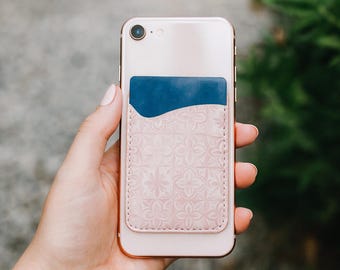 May include: A rose gold smartphone with a pink leather card holder attached to the back. The card holder has a blue pocket and a floral embossed design. The phone is held in a hand against a blurred green background.