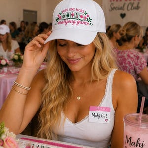 May include: A white baseball cap featuring the text "JUST ONE MORE mahjong game" in pink and green, accompanied by a graphic of four mahjong tiles, green leaves, and small pink flowers centered on the front panel of the hat.