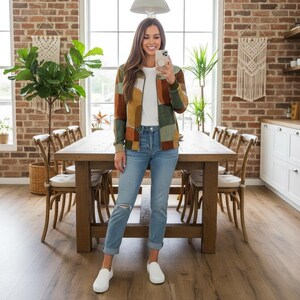 May include: A woman wearing a patchwork zip-up jacket in shades of green, brown, and yellow, paired with light blue jeans and white sneakers. The jacket has a ribbed collar and cuffs. The background includes a wooden dining table and chairs.