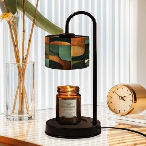 May include: A black metal candle warmer with a geometric patterned lampshade in shades of green, brown, and gold. A lit candle in a glass jar sits below. A gold-colored clock is visible in the background.
