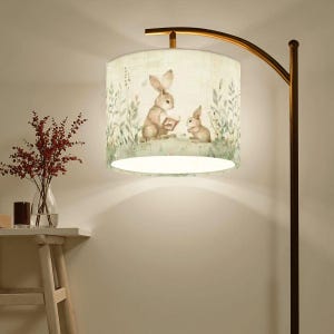 May include: A floor lamp with a lampshade featuring a watercolor illustration of two rabbits reading a book in a field of greenery. The lamp has a curved, bronze-colored arm and a dark brown pole. A vase of red flowers sits on a small table.