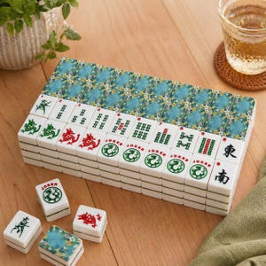 May include: A complete Mahjong set with white tiles featuring green and red symbols, including dragons and jokers. The set includes a floral patterned box and several loose tiles. The game is set on a wooden surface.