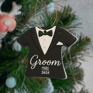 May include: A black and white tuxedo-shaped Christmas ornament with the word "Groom" in cursive script. The ornament includes the name "Mark" and the year "2025". It hangs from a gold string.
