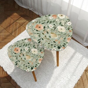 May include: Two nesting side tables with a floral design. The tables have a light green background with peach and white flowers and green leaves. The tables have wooden legs.