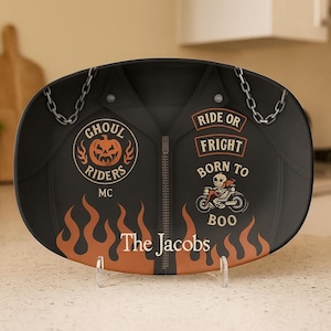 May include: Black oval decorative plate with a motorcycle jacket design. The plate features the text "Ghoul Riders MC", "Ride or Fright Born to Boo", and "The Jacobs". Orange flames and chain accents complete the design.