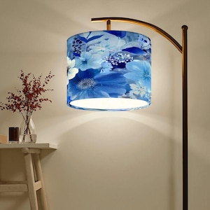May include: A floor lamp with a curved, bronze-colored metal frame. The lampshade is cylindrical and features a blue floral pattern with white and light blue flowers. The lamp is turned on, illuminating the room.