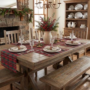 May include: A rustic wooden dining table set for a holiday meal. The table is adorned with red and green plaid placemats, white plates, and linen napkins. A centerpiece of greenery and berries sits in a cream-colored pitcher. Candles and glassware complete the festive setting.