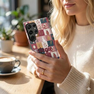 May include: A patterned phone case with a patchwork design featuring floral, bunny, and pineapple motifs in pink, blue, and white. The name "Angie" is written across the bottom. The phone case is held by a person.