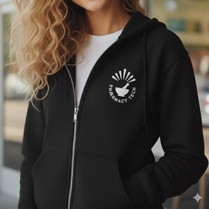 Puede incluir: Sudadera negra con cremallera y un gráfico blanco en la parte superior izquierda del pecho. El gráfico presenta un mortero y una maja con las palabras "PHARMACY TECH" en un diseño circular. La sudadera tiene una cremallera blanca.