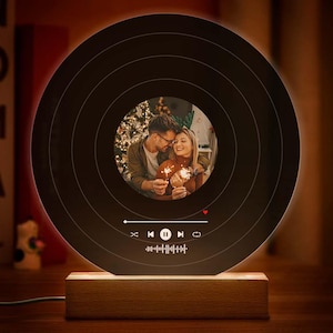 May include: A black circular lamp with a wooden base. The lamp features a round cutout in the center that displays a photo of a couple holding sparklers. The lamp has a music player interface with play, pause, and skip buttons.
