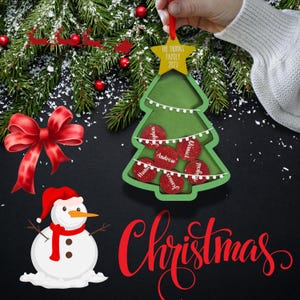 May include: A green Christmas tree ornament with a star-shaped top that reads "The Thomas Family 2023." The tree has red circular accents with names. A red bow and snowman are also in the image, along with the word "Christmas."