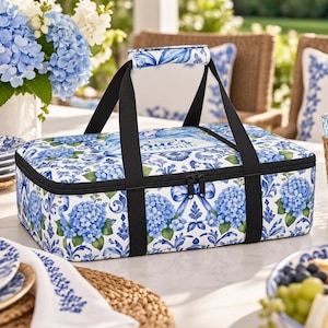 May include: A rectangular insulated food carrier featuring a blue and white floral hydrangea pattern with a central label reading Sue's Kitchen. It has black nylon straps, a padded handle, and a zippered closure, designed for transporting dishes.