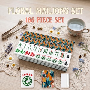 May include: A floral Mahjong set with 166 pieces. The tiles are white with green, red, and black symbols. The set includes a decorative box with a colorful floral pattern. A cup of tea and decorative elements are also present.