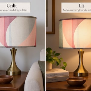 May include: A table lamp with a cylindrical fabric shade featuring an abstract pattern of pink, sage green, and grey curved shapes. The lamp has a metallic gold-toned hourglass-shaped base. The image shows the lamp in unlit and lit states.