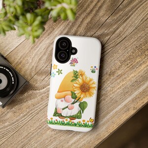 May include: White phone case with a cartoon gnome holding a sunflower. The gnome is wearing a yellow hat and a green shirt. The background is white with small flowers and mushrooms.