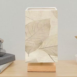 May include: A rectangular table lamp with a beige linen shade featuring a leaf pattern. The lamp sits on a natural wood base. The shade has a white interior, and the overall design is neutral and modern.