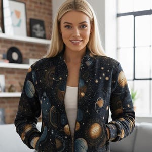 May include: A dark blue bomber jacket featuring a celestial design with planets and stars in shades of gold, blue, and brown. The jacket has a zip-up front and pockets. The model is wearing a white top underneath.