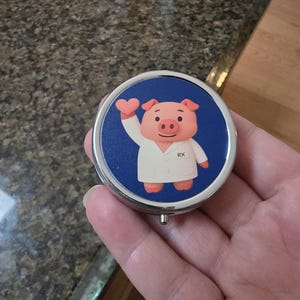 Puede incluir: Un pastillero redondo y plateado con fondo azul. Presenta un cerdo de dibujos animados con bata de laboratorio sosteniendo un coraz&oacute;n, con las letras "RX" en la bata. El pastillero se sostiene en una mano.