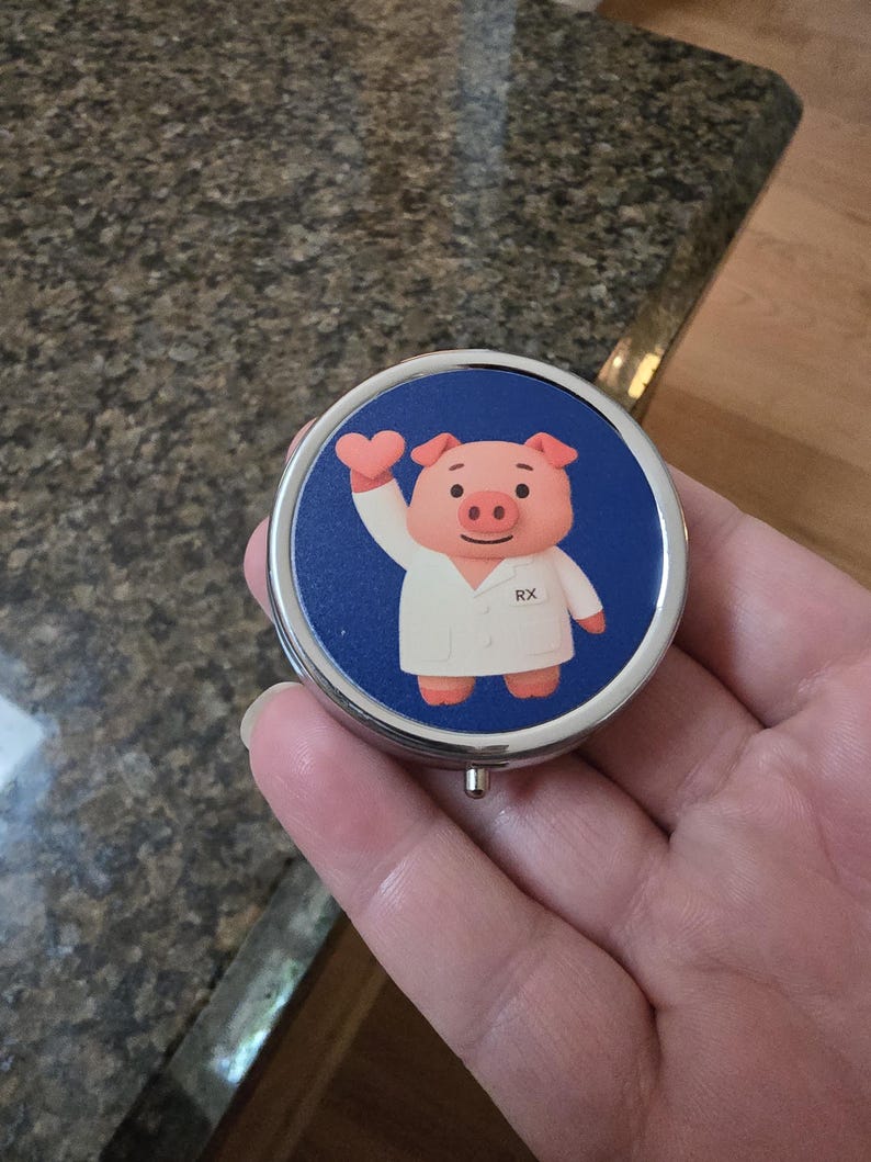 Puede incluir: Un pastillero redondo y plateado con fondo azul. Presenta un cerdo de dibujos animados con bata de laboratorio sosteniendo un coraz&oacute;n, con las letras "RX" en la bata. El pastillero se sostiene en una mano.
