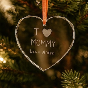May include: Clear, heart-shaped crystal ornament with the words "I ♥ MOMMY Love Aiden" etched on the surface. The ornament hangs from an orange ribbon against a blurred background of a Christmas tree.