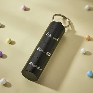 May include: Black pill organizer with a silver keychain. The organizer has four compartments labeled "Folic acid", "Vitamin B12", and "Amoxicillin".