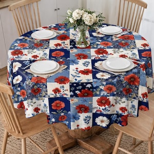 May include: A round tablecloth with a patchwork design featuring red, white, and blue floral patterns. The table is set with white plates, placemats, and a vase of white flowers. Wooden chairs surround the table, creating a dining setting.