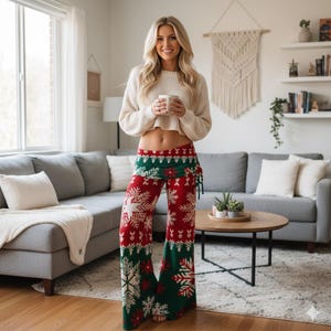 May include: Red and green Christmas-themed palazzo pants with a snowflake and reindeer pattern. The pants have a wide leg and a drawstring waist. The model is wearing a cream-colored sweater and holding a mug.
