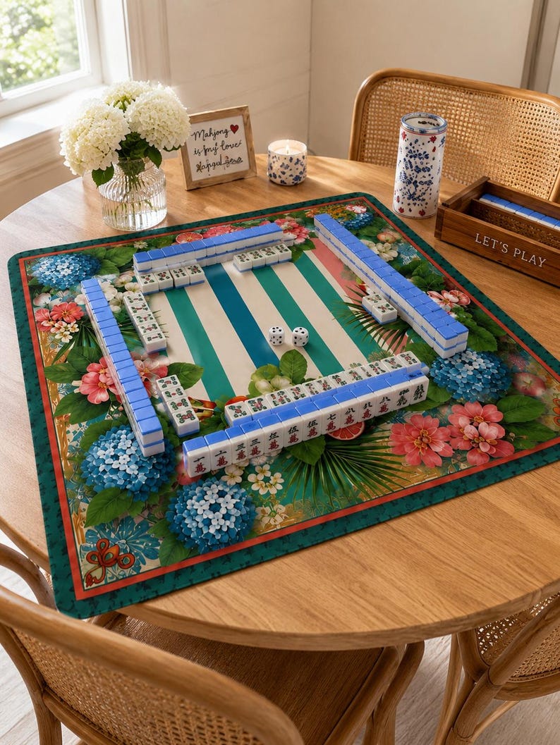 May include: A Mahjong game set on a round wooden table. The game board features a green and white striped design with blue and white tiles. The board is surrounded by floral patterns with blue and pink flowers. A wooden box with the words "LET'S PLAY" is visible.