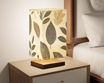 May include: A square-shaped table lamp with a wooden base and a cream-colored shade with a brown leaf pattern. The lamp is on a wooden nightstand with two drawers.