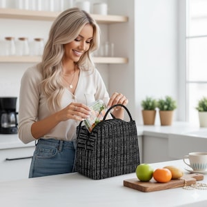 May include: A black and white patterned lunch bag with a top handle. The bag is open, and a clear container of food is being placed inside. A woman is smiling while holding the container. A wooden cutting board with fruit is in the foreground.