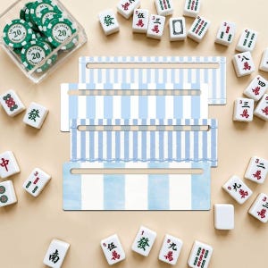 May include: Four rectangular wooden tile racks featuring a light blue and white vertical striped pattern. Each rack has a horizontal slot cut into the center. The racks are arranged in a stack, surrounded by scattered mahjong tiles and poker chips.