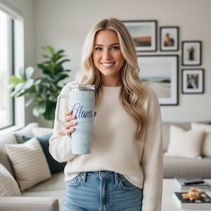May include: A light blue tumbler with a handle, featuring a floral pattern and the name "Alana" in script. The tumbler has a silver lid and is held by a person wearing a cream-colored sweater and blue jeans.
