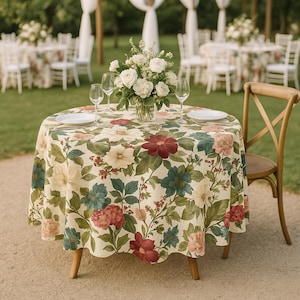 May include: A round table set for an outdoor event, covered with a floral tablecloth featuring burgundy, blue, and cream-colored flowers. A vase of white roses sits in the center, with plates and wine glasses. A wooden chair is positioned to the side.