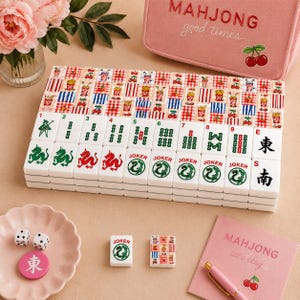 May include: A set of mahjong tiles featuring white tiles with green and red symbols, including dragons and jokers, alongside tiles with red and blue checkered patterns. Includes two white dice, a pink storage case, and a pink notebook with text.