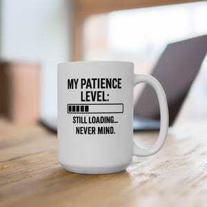 May include: White ceramic mug with the text "MY PATIENCE LEVEL: Still Loading... Never Mind." The mug has a black graphic of a loading bar. The mug is on a wooden surface with a laptop in the background.