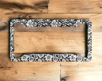 May include: Black and white floral patterned license plate frame. The frame is made of metal and has a glossy finish.
