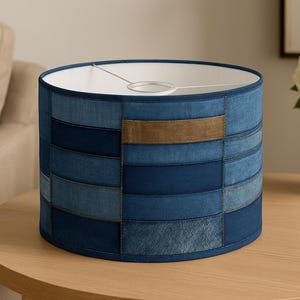 May include: A cylindrical lampshade with a patchwork design made from various shades of blue denim. A rectangular patch of brown fabric is centered on the shade. The lampshade sits on a round wooden table.