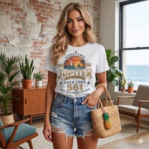 Könnte beinhalten: Weißes T-Shirt mit einem Aufdruck mit Leuchtturm, Sonnenuntergang und dem Text "Hometown Jupiter Florida Area Code 561". Die Person trägt Jeansshorts und eine Korbtasche mit Ananas-Anhänger.
