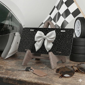 May include: A black license plate with a leopard print pattern and a large white bow. The plate is displayed on a wooden stand, with a racing flag and tires in the background. Sunglasses and keys are on the wooden surface.