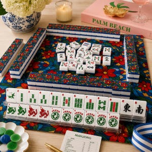 Puede incluir: Un juego de Mahjong completo con fichas blancas con s&iacute;mbolos verdes y rojos, presentado sobre un tapete con estampado floral. El juego incluye estantes, dados y una hoja de puntuaci&oacute;n.