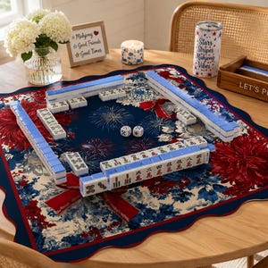May include: A Mahjong set arranged on a navy blue and red floral patterned mat. The set includes blue and white tiles, dice, and a wooden box that says "LET'S PLAY". A candle and a tumbler with the text "Stars Stripes Mahjong Vibes" are also present.