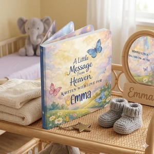 Puede incluir: Un libro para bebé personalizado, titulado "A Little Message From Heaven", con el nombre "Emma" en la portada. El libro presenta ilustraciones de mariposas y se muestra con botines de bebé de ganchillo y un dije en forma de estrella.