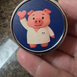 Puede incluir: Espejo compacto redondo con borde plateado, con un dise&ntilde;o de cerdo de dibujos animados con bata blanca con "RX" impreso. El cerdo rosa levanta una mano en forma de coraz&oacute;n sobre un fondo azul oscuro.