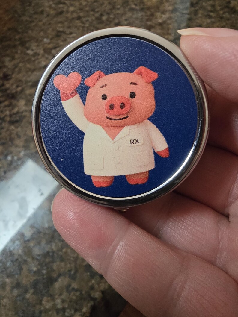 Puede incluir: Espejo compacto redondo con borde plateado, con un dise&ntilde;o de cerdo de dibujos animados con bata blanca con "RX" impreso. El cerdo rosa levanta una mano en forma de coraz&oacute;n sobre un fondo azul oscuro.