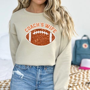 May include: A tan sweatshirt with the text "COACH'S WIFE" in orange letters with a glitter football graphic.