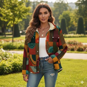 May include: A colorful, patterned jacket with a cream-colored collar and a white interior. The jacket features a geometric design in shades of red, orange, green, and yellow. The person is wearing a white top and blue jeans. The background is a park with green grass and trees.